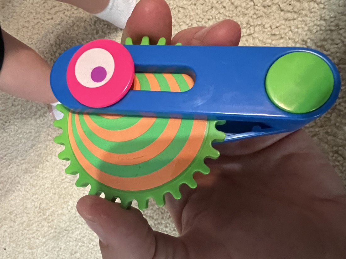Front photo of a colorful child's toy with a green gear, blue handle, and pink cap